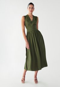 Vestido midi verde oliva con escote en V, sin mangas, cintura ajustada y falda plisada. Combinado con unos tacones blancos de tiras.