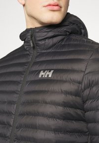 Černá zateplená bunda s kapucí, vybavená předním zipem a horizontálně prošívaným designem. Logo "HH" zobrazené šedě na hrudi.