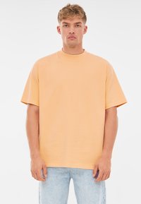 Korte mouwen, ronde hals t-shirt van lichtoranje katoen. Heeft een relaxte pasvorm en een soepele textuur, gecombineerd met blauwe spijkerbroeken.