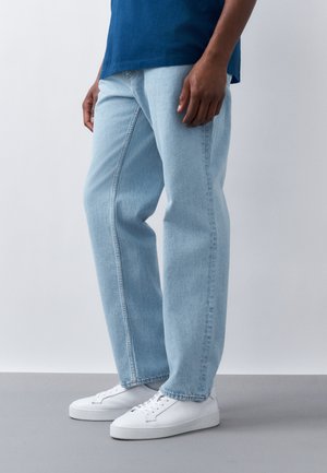 Osoba oblečená v svetlomodrých džínsach s rovnými nohavicami, modrej košeli a bielych šnurovacích teniskách stojaca na sivom podlaží.