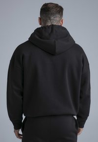 Zwarte katoenen hoodie met een losse pasvorm en een grote capuchon, voorzien van afgezakte schouders en een gladde textuur. Zichtbare stiksel-details.