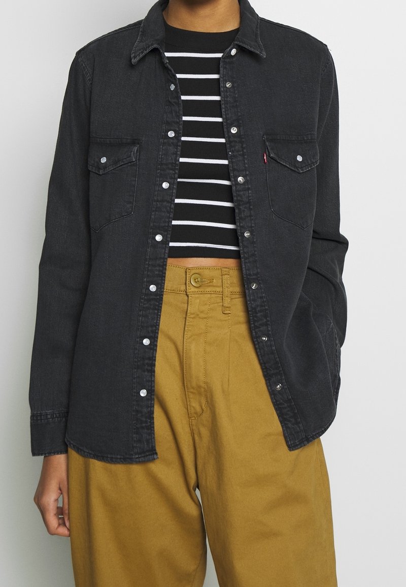 Camicia di jeans nera con bottoni a pressione, due tasche sul petto e design con colletto, abbinata a pantaloni beige a vita alta e a un top a righe bianco e nere.