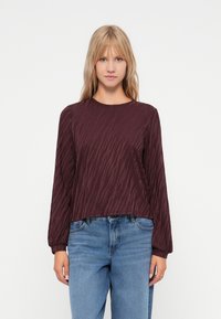 Burgunderfarbenes Langarmoberteil mit strukturiertem Wellenmuster, rundem Ausschnitt und lässiger Passform, kombiniert mit hellblauen Jeans.