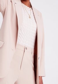 Blazer rose clair en tissu lisse, avec une fermeture à un bouton et des poches avant, associé à un haut blanc texturé.