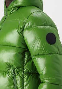 Zeleni puffer jakna s sjajnom završnom obradom, kvadratnom teksturom, zatvorenim džepom i okruglim logotipom na gornjoj ruci.