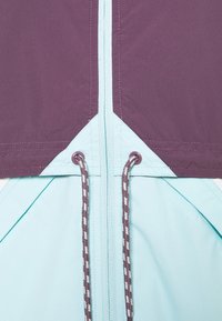 Rompevientos con bloques de color, con una sección superior de púrpura oscuro, una sección inferior de azul claro, cremallera frontal y detalles de cordón en marrón y blanco.