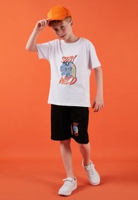 Garçon portant une casquette orange, un t-shirt blanc "Stay Wild" avec un motif de dinosaure, un short noir avec le même motif, et des baskets blanches sur fond orange.