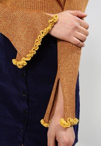 Sweter w kolorze rdzy o prążkowanej fakturze z dzwonkowatymi rękawami ozdobionymi żółtą falbaną; do tego ciemnoniebieska spódnica ze sztruksu z guzikami z przodu.