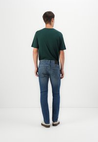 T-shirt vert à manches courtes, jeans slim-fit bleus et baskets claires. Design minimaliste, uni, avec une étiquette visible sur la ceinture.