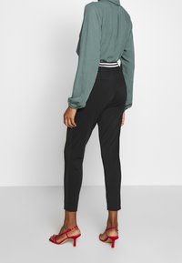 Grön texturerad blus med krage, svarta figurnära byxor och röda sandaletter med remmar. Noterbara detaljer inkluderar en kontrasterande midjeband och smal passform.