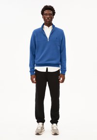 Sweatshirt zippé bleu avec col montant, porté par-dessus une chemise blanche à boutons. Associé à un pantalon noir et des baskets grises.