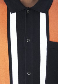 Gros plan d'une chemise en maille noire avec des rayures blanches verticales, des panneaux latéraux marron, un col noir, des boutons-pression en métal et une poche poitrine.