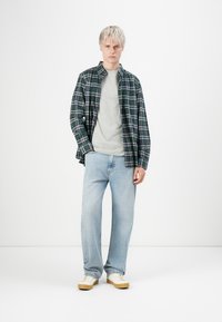 Jong persoon staat in een groen geruit shirt over een grijs sweatshirt, lichte spijkerbroek en witte sneakers met rubberen zolen.
