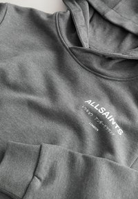 Sweat à capuche gris avec poignets côtelés, arborant le texte "ALLSAINTS" ainsi que des caractères japonais et "LONDON" imprimés sur la poitrine.