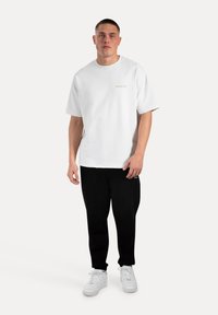 Weißes Baumwoll-T-Shirt mit Rundhalsausschnitt und kurzen Ärmeln, ausgestattet mit einem kleinen goldenen Logo auf der Brust. Kombiniert mit schwarzen Sweatpants und weißen Turnschuhen.