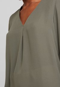 Olivengrüne Bluse mit V-Ausschnitt und lockerer Passform, die eine weiche, glatte Textur und subtile Faltendetails an der Vorderseite aufweist.