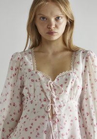 Blusa a fantasia floreale in tessuto leggero con maniche a sbuffo, профili in pizzo, scollatura profonda e dettaglio con coulisse in vita. Sfumature di rosa tenue predominano.