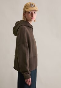 Braunes Kapuzensweatshirt mit lockerem Sitz, kombiniert mit einer hellbeigen Kappe mit großem numerischem Design. Glatte Textur, minimalistischer Stil.
