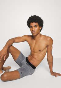 Sivi športni kratki hlači s črnim pasom in majhno logotipom; prilegajoč kroj in gladka tekstura, nošeni s strani osebe, ki sedi na površini.