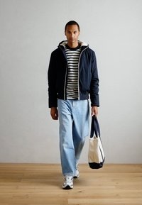 Marineisoleret jakke, stribet skjorte, lyseblå jeans og hvide sneakers. Holder en canvas taske med marine accenter. Trægulv i baggrunden.