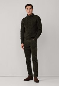 Man med mörkgrön kabelstickad polotröja, matchande byxor och bruna mockastövlar, stående mot en enfärgad ljus vägg.