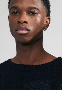 Pull en tricot texturé bleu marine avec un large col, porté par une personne aux cheveux courts et à une boucle d'oreille en or. La peau semble légèrement brillante.