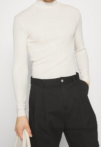 Jersey de manga larga color beige claro con cuello alto, hecho de tela acanalada, combinado con pantalones negros de tiro alto que presentan un cierre frontal con botón.