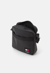 Czarna syntetyczna torba na ramię z zamkiem błyskawicznym, przednią kieszenią i plecionym paskiem; charakteryzuje się naszywką z logo i teksturowaną powierzchnią.