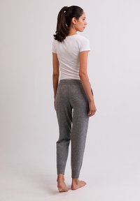 Graue Fleece-Jogginghosen mit einem eng anliegenden Bund und elastischem Taillenbund, kombiniert mit einem weißen, eng anliegenden T-Shirt. Das Modell steht barfuß und blickt von der Kamera weg.