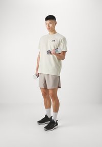 Šviesiai smėlinis marškinėliai su Under Armour logotipu, šviesiai pilkos šortai, juodi sportiniai batai ir sidabriniai hanteliai kiekvienoje rankoje.