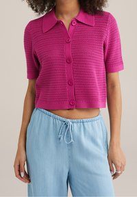 Fuchsia, stickad kortärmad cardigan med krage, knappsl Closure och strukturerat mönster. Matchas med ljusblå byxor med dragsko i midjan.