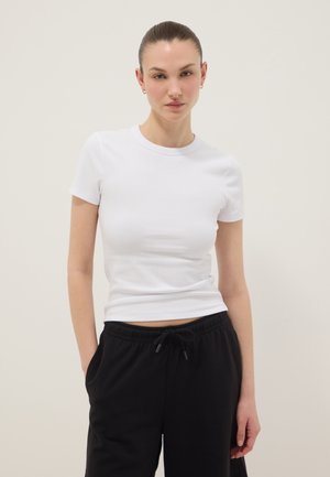 Donna con capelli raccolti indossa piccoli orecchini a cerchio, una maglietta bianca aderente a maniche corte e pantaloni neri con coulisse, in piedi davanti a uno sfondo uniforme.