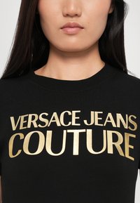 Melns kokvilnas T-krekls ar izteiksmīgiem zeltainiem burtiem, kas uzrakstīti "VERSACE JEANS COUTURE." Vienkāršs dizains ar klasisku apaļu kakla izgriezumu.