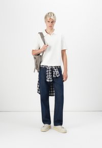 White polo shirt, dark blue jeans, and a plaid shirt tied at the waist. Beige crossbody bag and white sneakers complete the look.