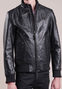 Veste bomber en cuir noir avec fermeture éclair à l'avant, poignets et ourlet côtelés, et poches latérales. Inclut un petit patch sur le haut de la poitrine.