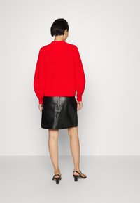 Femme debout, vue de dos, portant un pull rouge vif, une jupe en cuir noire avec une fermeture éclair, et des sandales à talons noires sur un fond clair.