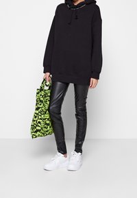 Persona che indossa un ampio hoodie nero, pantaloni di pelle neri, sneakers bianche, mentre tiene una borsa a spalla verde e nera con motivo.