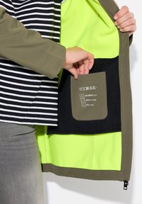 L'interno della giacca impermeabile presenta un tessuto di un vivace verde neon e una tasca nera etichettata con le istruzioni per la cura. Design esterno a righe, verde oliva.