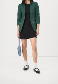 Zelený blazer s kapsami na zip přes černé šaty, doplněný černými botami, bílými ponožkami a světle modrou kabelkou.