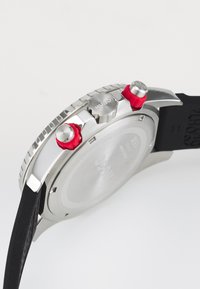 Silver stainless steel watch with a black rubber strap, featuring two red crown accents and a textured bezel. Back is smooth and engraved.