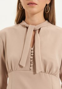 Blusa beige con nodo al collo, caratterizzata da un scollo a fessura, tessuto morbido e bottoni decorativi sul davanti. Maniche lunghe con una vestibilità comoda.
