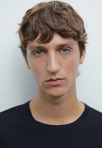 Ritratto di un giovane con capelli castani chiari spettinati, che indossa una semplice maglietta nera. Ha occhi blu-verdi e un'espressione neutra.