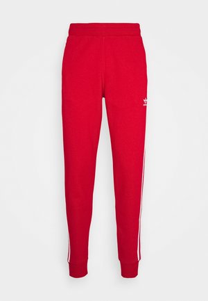Pantalon de survêtement rouge Adidas avec bandes latérales blanches, taille et poignets élastiques, présenté sur fond blanc uni.