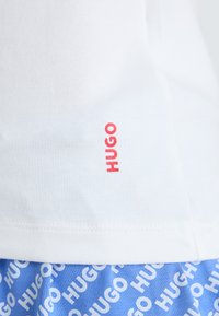 Balta medvilninė marškinėliai turi raudoną "HUGO" logotipą šone. Po jais, mėlyni šortai rodo pakartotiną baltą "HUGO" tekstą.