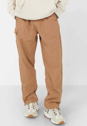 Tan cargobroek met een ontspannen pasvorm, voorzien van zijzakken, knopen op de mouwen en een gladde stofstructuur. Gecombineerd met witte sneakers.