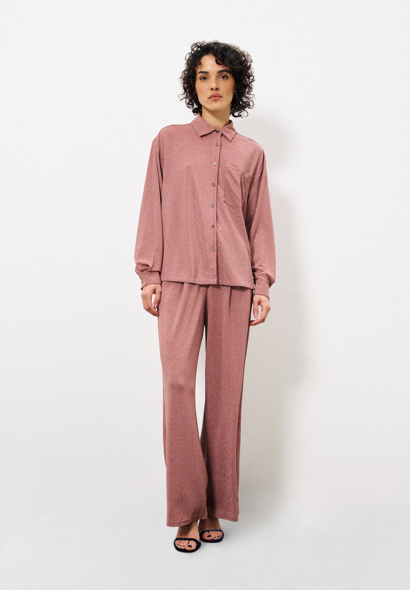 Helles rosa Button-Up-Bluse mit einer Brusttasche, kombiniert mit passenden weit geschnittenen Hosen, beide aus einem weichen, strukturierten Stoff gefertigt.