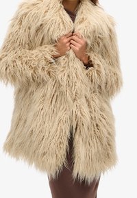 Beige kunstpälsjacka med lång, rufsig textur, bred krage och öppen fram. Bärs över en brun klänning, utan synliga knäppningar.