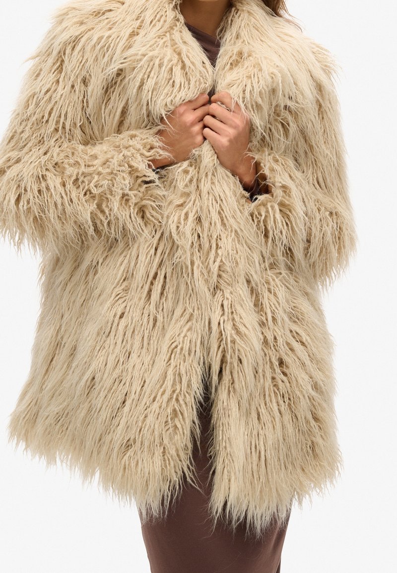 Beige kunstpälsjacka med lång, rufsig textur, bred krage och öppen fram. Bärs över en brun klänning, utan synliga knäppningar.