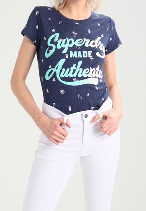 Kobieta w granatowym t-shircie z napisem "Superdry Made Authentic" i małymi białymi zdobieniami, ubrana w białe, wysokowcięte jeansy, trzymająca pętle paska.