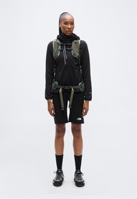 Μαύρο εξωτερικό μπουφάν με κουκούλα, πράσινες λουρίδες, σετ με μαύρο σορτς και αθλητικά παπούτσια. Απλός σχεδιασμός, λειτουργικό για υπαίθριες δραστηριότητες.